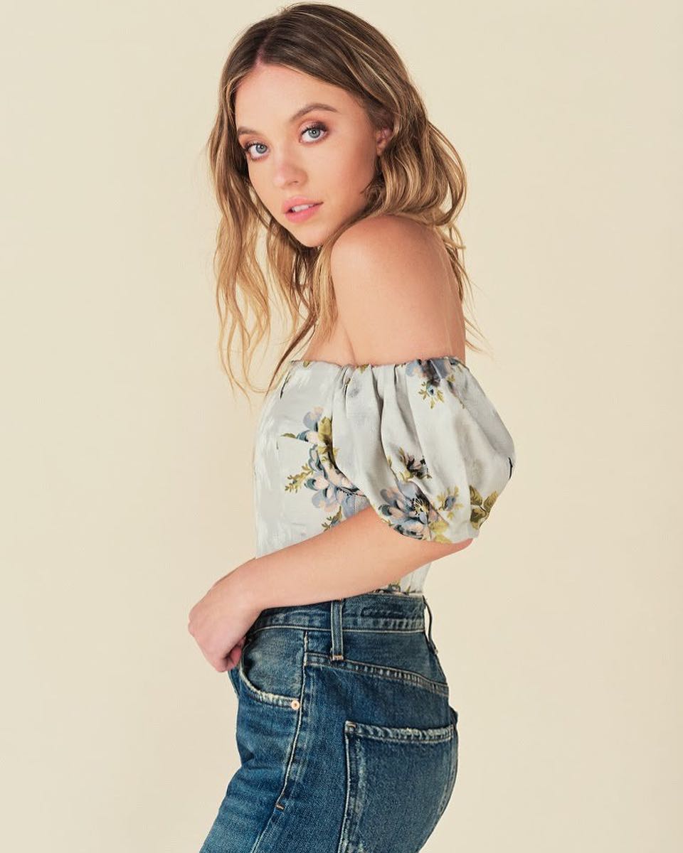 a side profile of a young blonde woman modeling a light blue floral offtheshoulder blouse with puff sleeves, paired with highwaisted dark wash denim jeans. she is posing against a light beige backdrop, her hair styled in loose waves. the overall style is feminine and romantic.