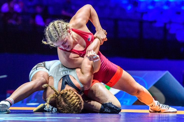 two female wrestlers are midmatch, with one on top and one on the bottom. the wrestler on top is in a red and white singlet, and the wrestler on the bottom is in a black and white singlet. the wrestler on top has a strong grip on the wrestler on the bottom's arm and is pushing her opponent down to the mat. they both have blonde hair and are wearing wrestling shoes. they are competing on a blue mat with a white mat border, and the background is blurred, showing the audience and other details of the wrestling competition.