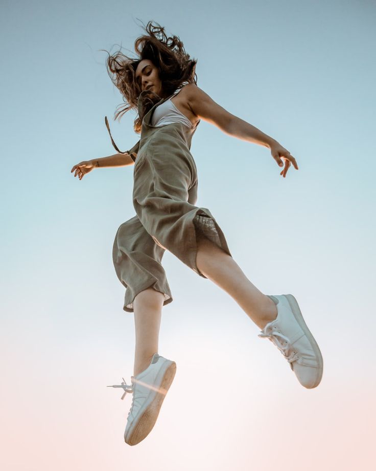 a young woman in a white tank top and green jumpsuit jumps with a carefree and joyful expression. her long brown hair is flowing in the air as she leaps high with both arms out to the sides. she is wearing white sneakers and the shot is taken from a low angle, looking up at her. the background is a bright blue sky with clouds and the image gives the feeling of summer and freedom.