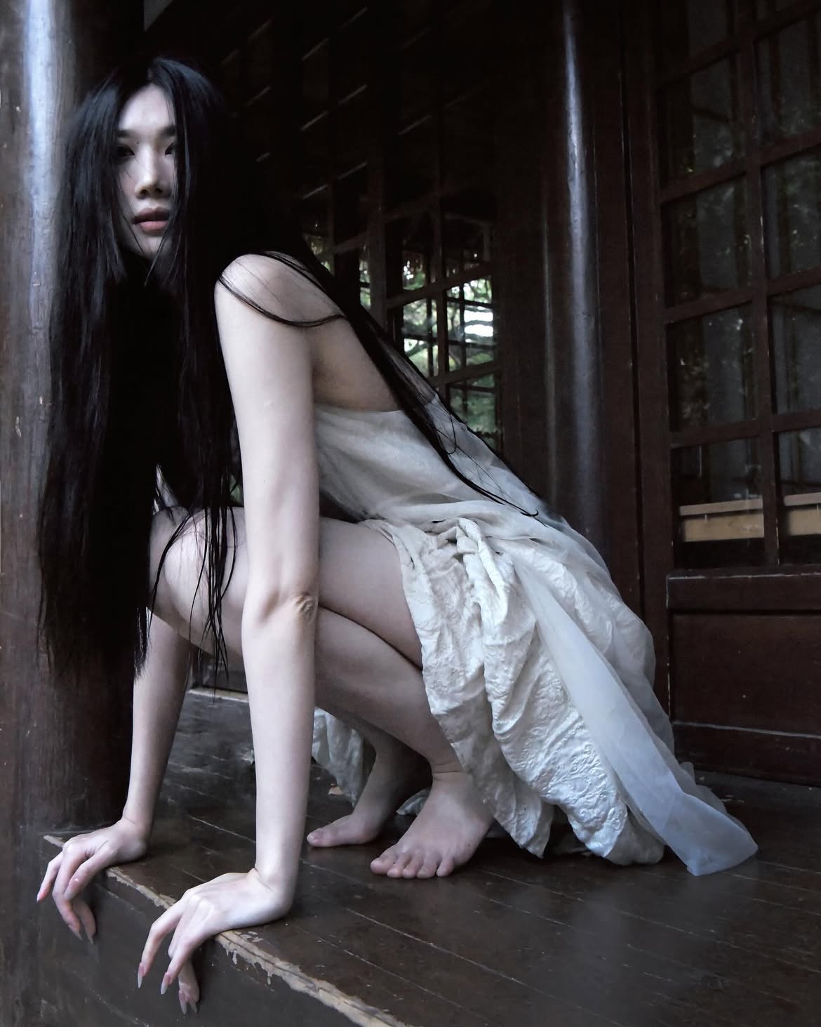 a woman with long, dark hair, wearing a white dress, is crouching on a wooden veranda. she is barefoot and has a spooky or ghostly appearance. the setting appears to be an old building with wooden windows. the overall mood is dark and mysterious.