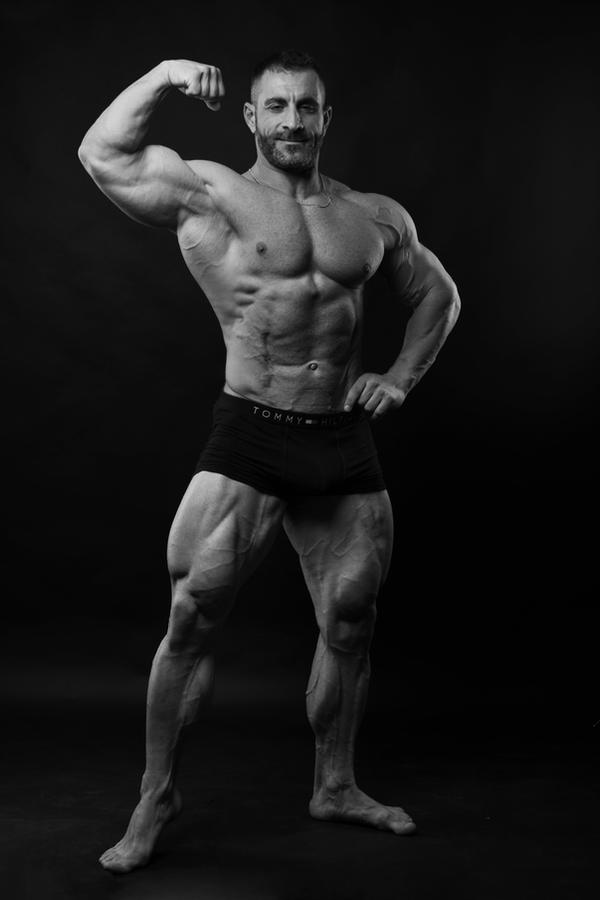 the image is a black and white studio shot of a muscular male bodybuilder. he is posing, flexing his right bicep, showing off his highly defined physique.  his abdominal muscles are clearly visible, along with prominent veins in his arms and legs. he is wearing black underwear with a visible tommy hilfiger logo. the background is a solid, dark color, which draws all attention to the subject's body. the overall mood is one of strength, health, and fitness.