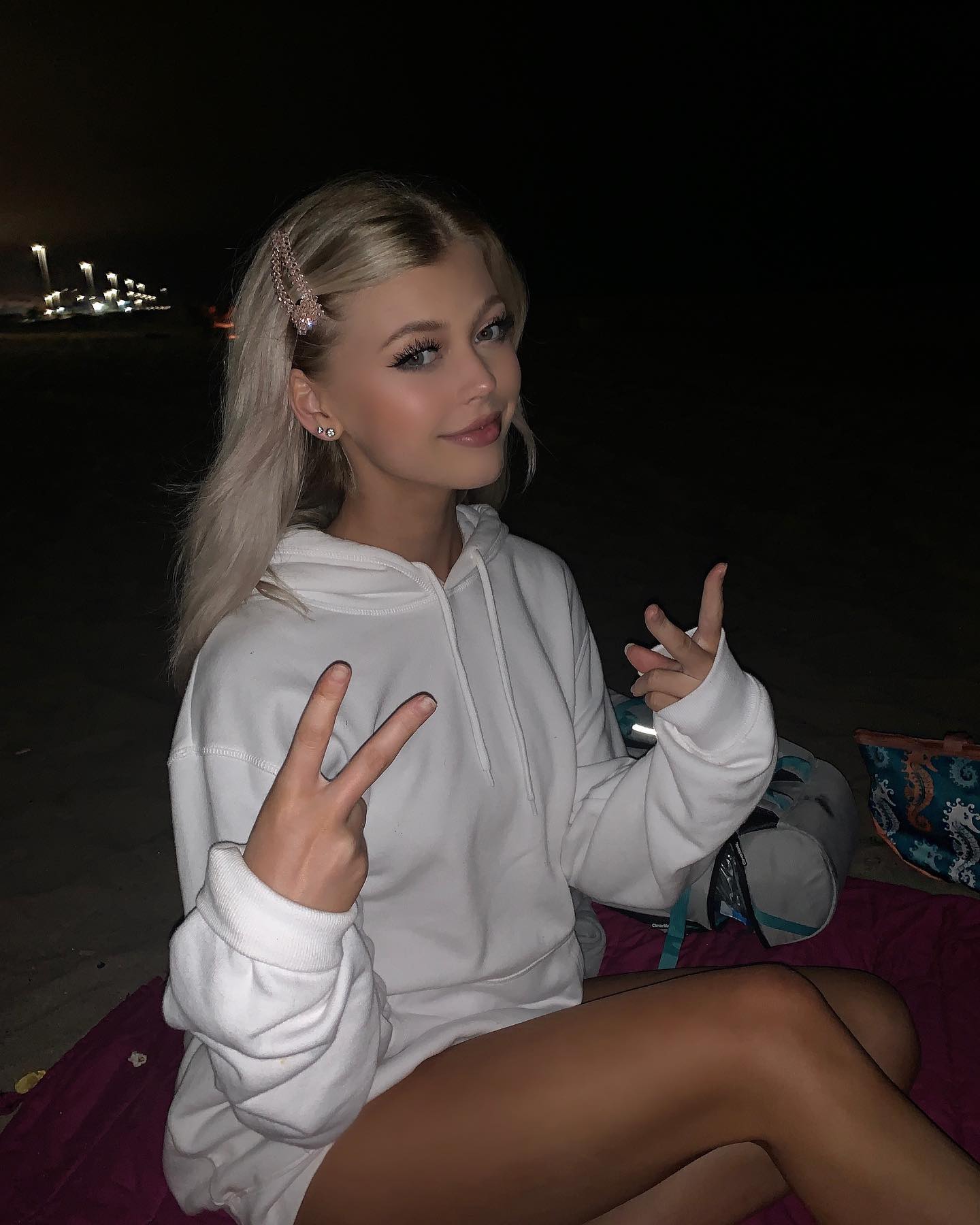 a blonde woman in a white hoodie is sitting on a beach at night. she is making a peace sign with both hands and wearing a hair clip and earrings. there is a backpack and a blanket behind her.