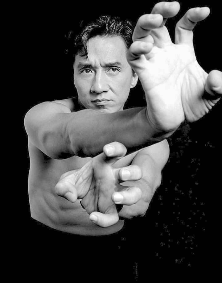 a black and white portrait of a man. he is looking at the camera with a serious expression. his hand is outstretched in front of him, as if he is about to strike. he is wearing a black shirt and his arms are bare. the image is welllit and the subject is in focus. he appears to be an actor or martial artist.