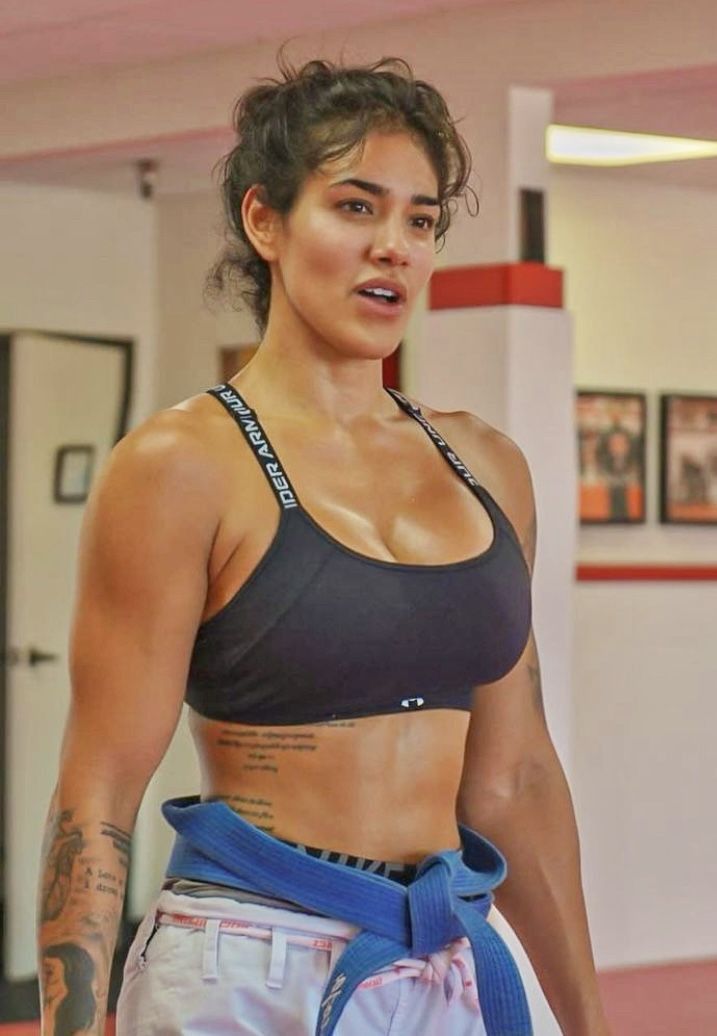a woman in a gym stands facing slightly to the left of the camera. she is wearing a black sports bra and blue jiujitsu pants with a blue belt. she is muscular and has a tattoo on her left rib cage and left arm. the background is a gym with white walls, red trim, and a door on the left side. there are also some framed pictures on the wall in the background. the lighting is bright and natural, coming from the overhead lights and possibly a window out of frame.