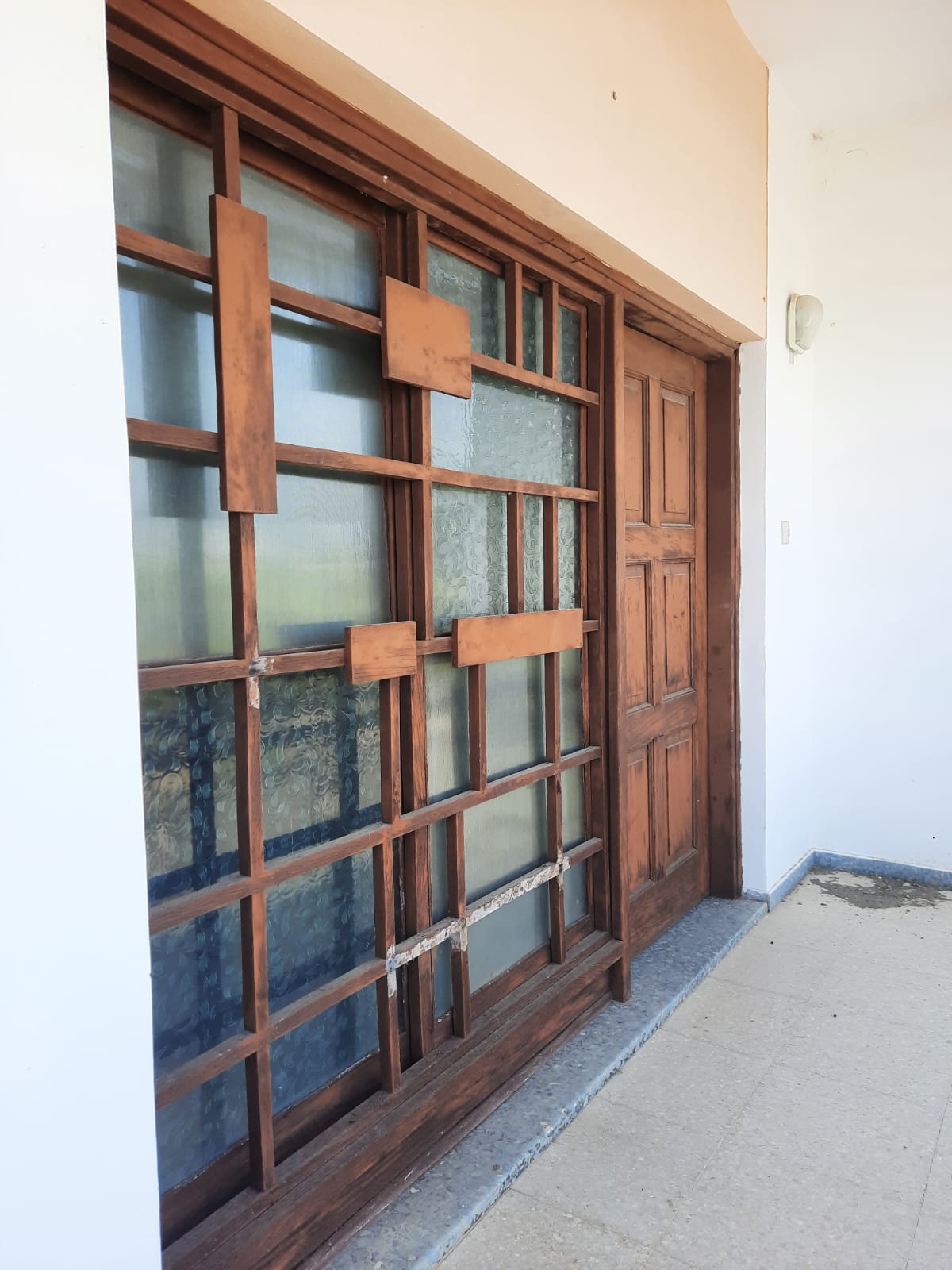 an exterior shot features a wooden door and a large window with a wooden grid frame. the door is positioned to the right of the window. the wooden frame around the window has a geometric design. the structures are part of a building's facade, with the setting appearing to be a porch area. the window has glass panes and the door is solid wood.