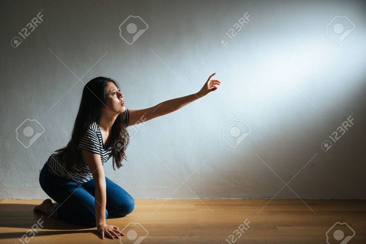 a young asian woman is kneeling on a wooden floor. she is reaching out with her right hand towards the light.  she is wearing jeans and a striped top.  she has long dark hair.  the background is a grey wall.  the woman appears to be hopeful, but also lost and desperate.
