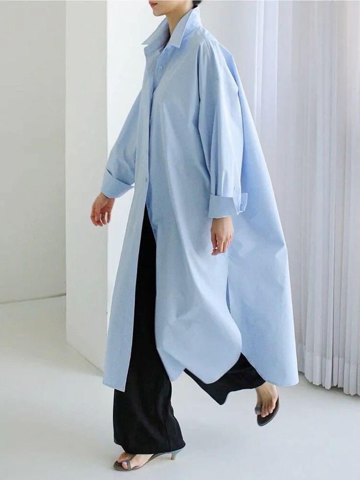 a woman wearing a long, light blue shirt dress with a white collar and buttons down the front. she is wearing black pants underneath the dress and has her hair pulled back away from her face. she is standing with one foot slightly in front of the other and is looking to the side. the background is white.