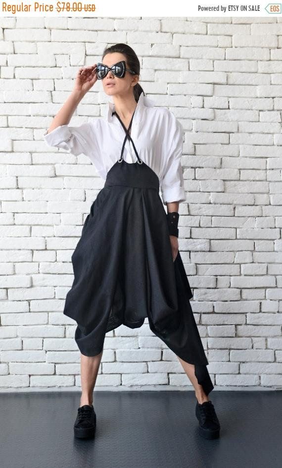 a woman wearing a black jumpsuit with a white shirt and black sunglasses is posing in front of a white brick wall. she is wearing black shoes.