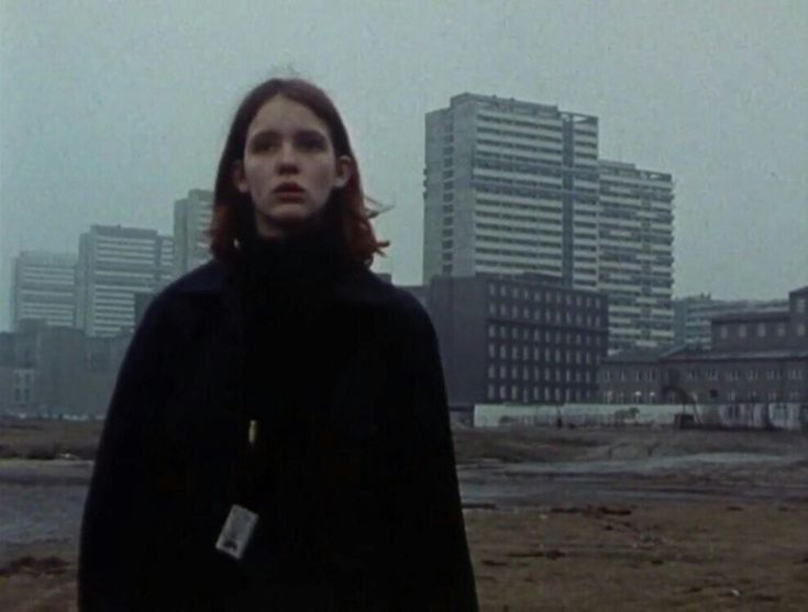 the image is a still from a film (likely from the 1970s), showing a young woman with shoulderlength reddishbrown hair. she's wearing a dark, long coat and stands in a barren, seemingly postindustrial landscape.  the background features a backdrop of imposing highrise apartment blocks, which dominate the scene. the overall mood is bleak and desolate, with a grey, overcast sky adding to the somber atmosphere. the woman's expression is serious and somewhat pensive, conveying a sense of solitude. the style of the image and clothing points towards a 1970s setting, possibly somewhere in the uk.