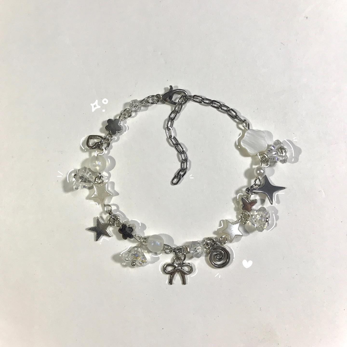 a closeup of a silver bracelet with various charms including stars, flowers, beads, a bow, and a spiral. the bracelet is made of a chain with an adjustable extension. the charms are silver, white, and clear.