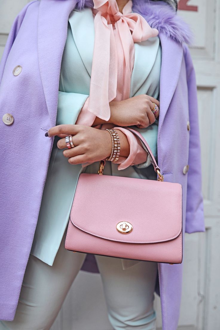loseup view of a woman wearing a stylish pastelcolored outfit. she is wearing a lavendercolored wool coat over a light mint green blazer, a pink silk bowtie, and white pants. she carries a pale pink satchel bag and has several gold rings and bracelets on her wrists. the setting appears to be outdoors during the day, with a blurry background suggesting a building or wall. the overall aesthetic is feminine, elegant, and springlike.