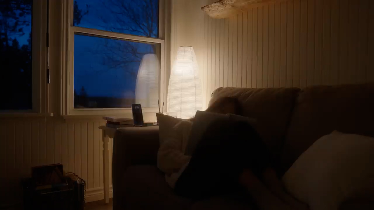 a woman is curled up on a couch in the evening, illuminated by a warm lamp. the room is dimly lit, with the only other light source coming from the window, which reveals a dark evening sky. the scene evokes a sense of peace and relaxation.