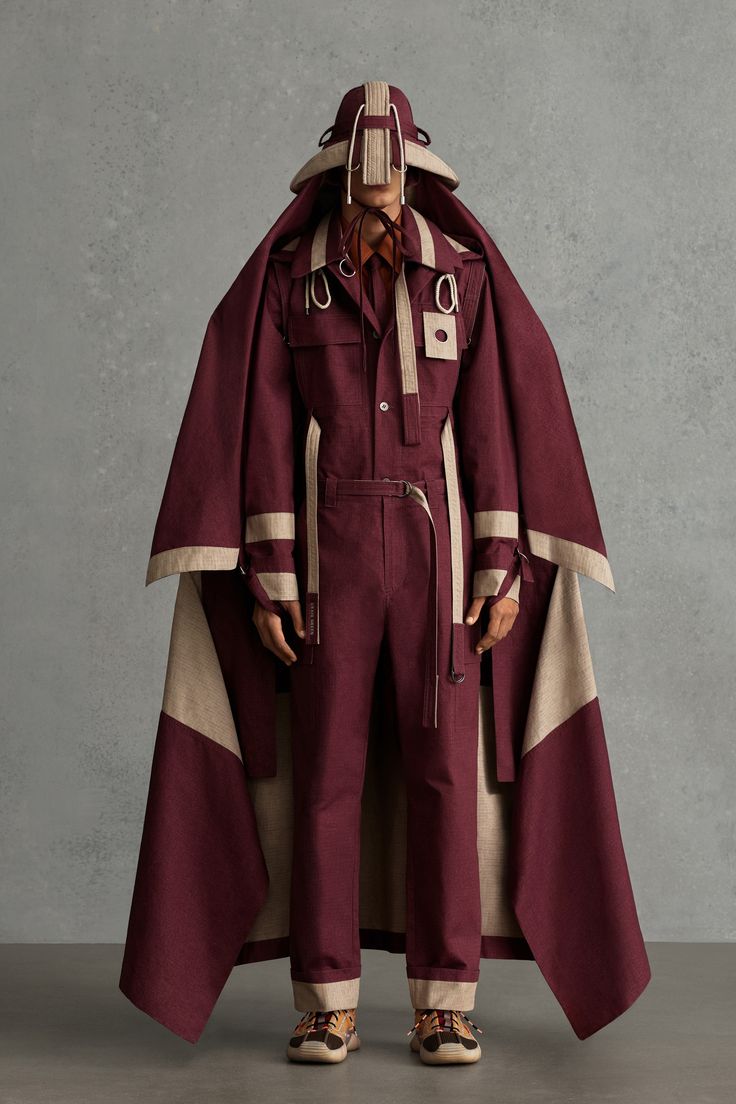 a fashion model stands against a grey backdrop wearing an allmaroon jumpsuit with a beige cape and hat. the cape is a mix of burgundy and beige fabrics, creating a layered effect. they're wearing brown sneakers.