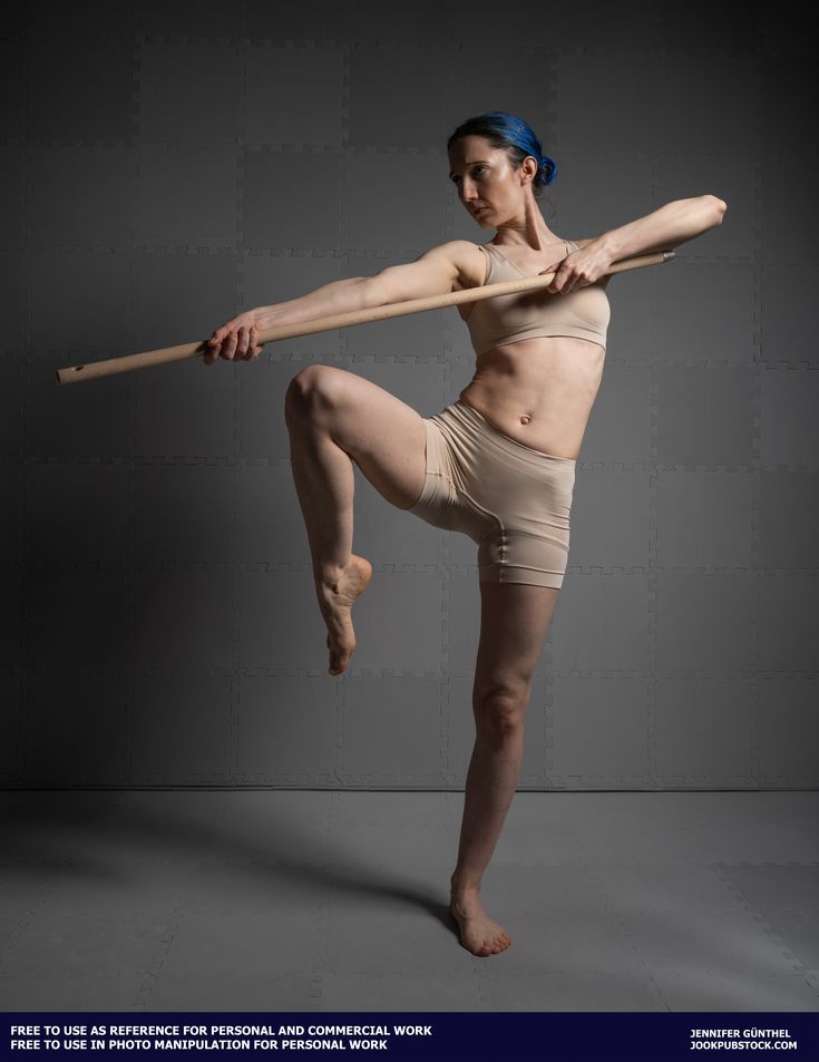 a full shot of a woman with blue hair, wearing a beige sports bra and shorts, holding a long wooden staff. she is in a dynamic pose, her body angled and her leg raised, suggesting martial arts or dance. the background is a simple grey studio backdrop.  the image is presented as reference material for personal and commercial use.