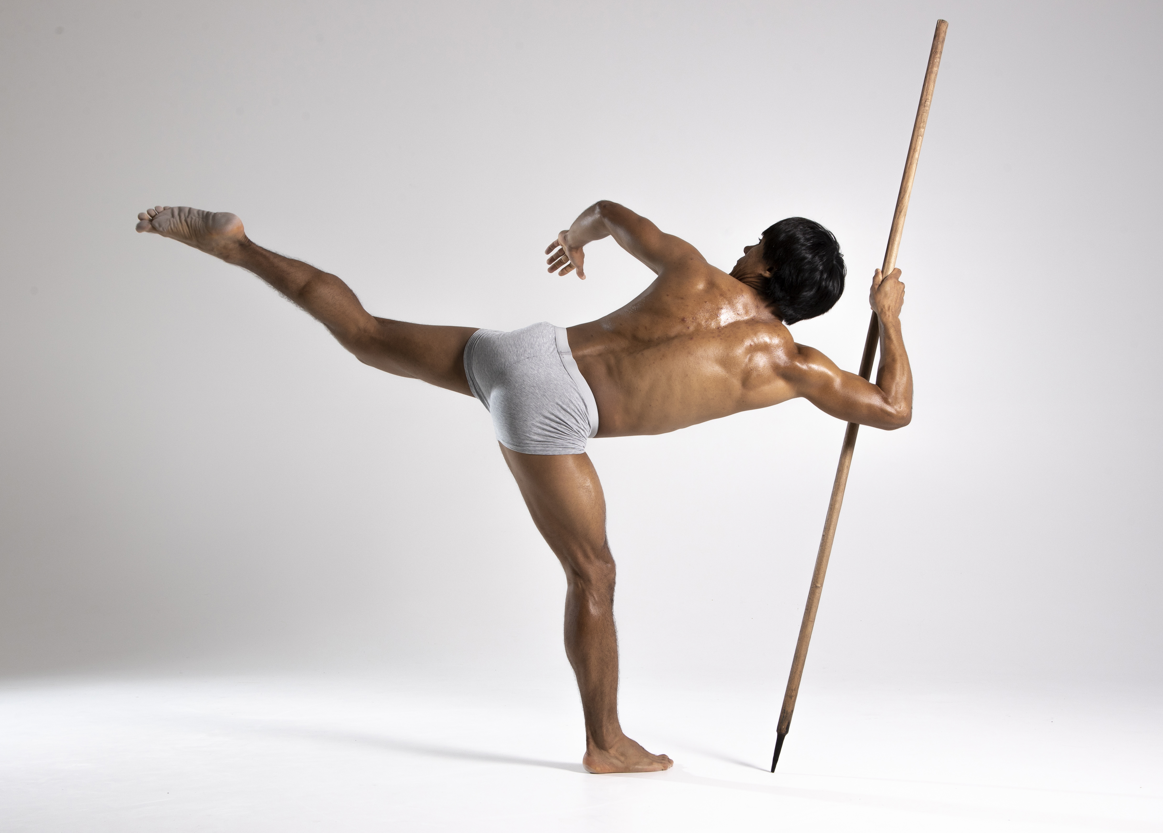 a shirtless man stands in a balancing pose with one leg extended behind him and the other leg planted on the ground. he holds a spear in his hands. he is barefoot. the background is white.