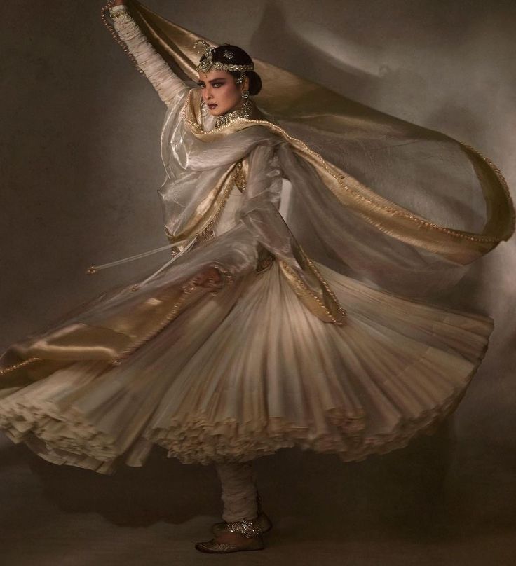 a woman in a traditional indian white dress with gold embroidery dances gracefully in a studio. her dress is swirling and flowing around her as she poses with elegance. she has a beautiful headpiece and jewelry on her head and neck. the background is a neutral beige color, allowing her to be the main focus of the photo.