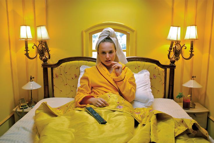 a woman in a yellow robe sits in a yellow bedroom. she's sitting up in bed with a white pillow and blanket and holding a tv remote. the bedroom is decorated in yellow and white with lamps on the nightstands. the woman looks relaxed and comfortable. the image is taken in a home setting, perhaps during a movie or film production.