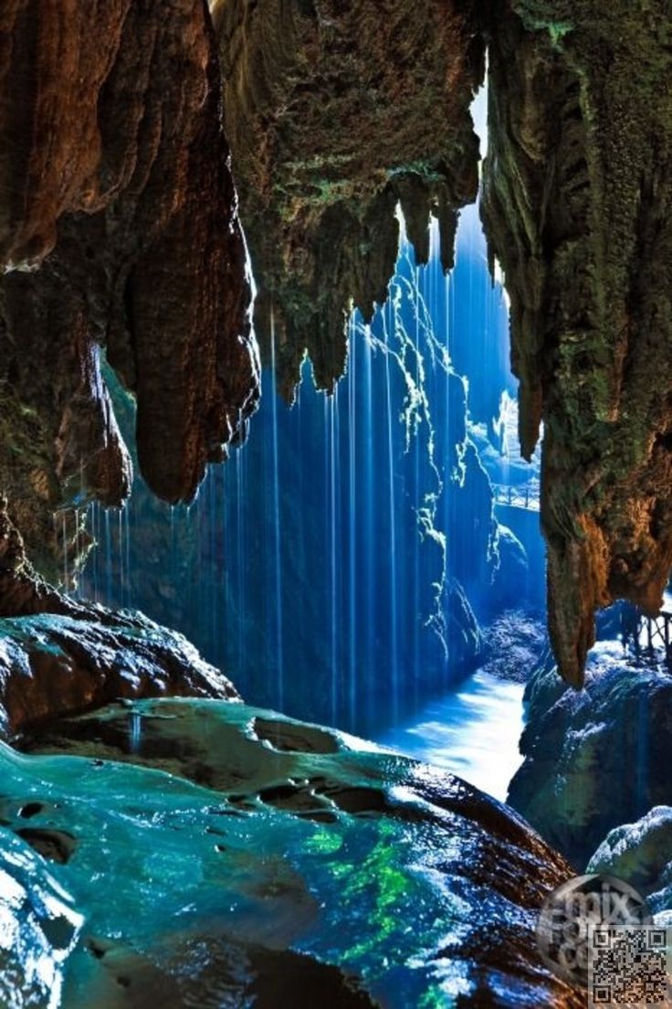 the image shows an underground cave with a river flowing through it. stalactites hang from the ceiling, and the water is turquoise in color. blue light illuminates the scene, creating a dramatic effect. a small bridge is visible in the distance.