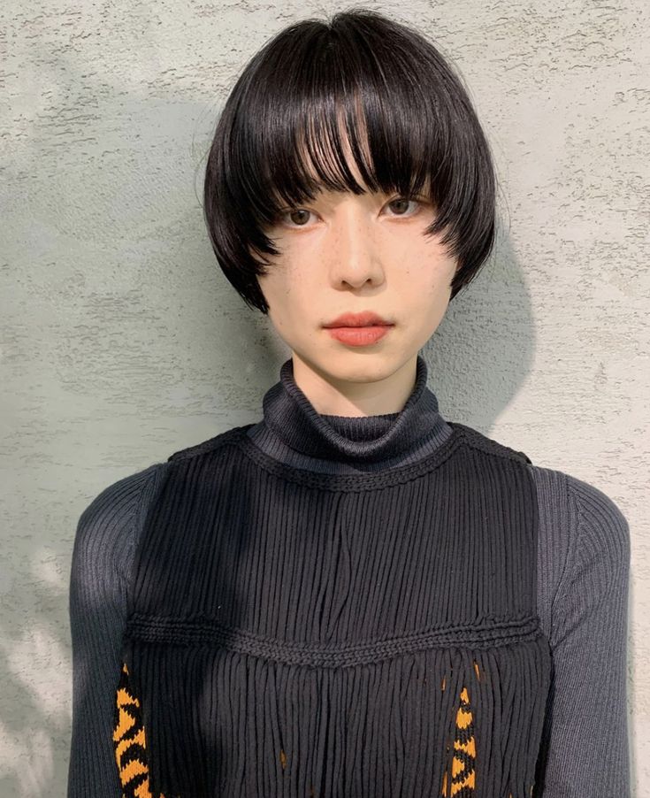 a woman with short black hair with a fringe, wearing a black turtleneck sweater with a black fringe detail. she is looking at the camera.