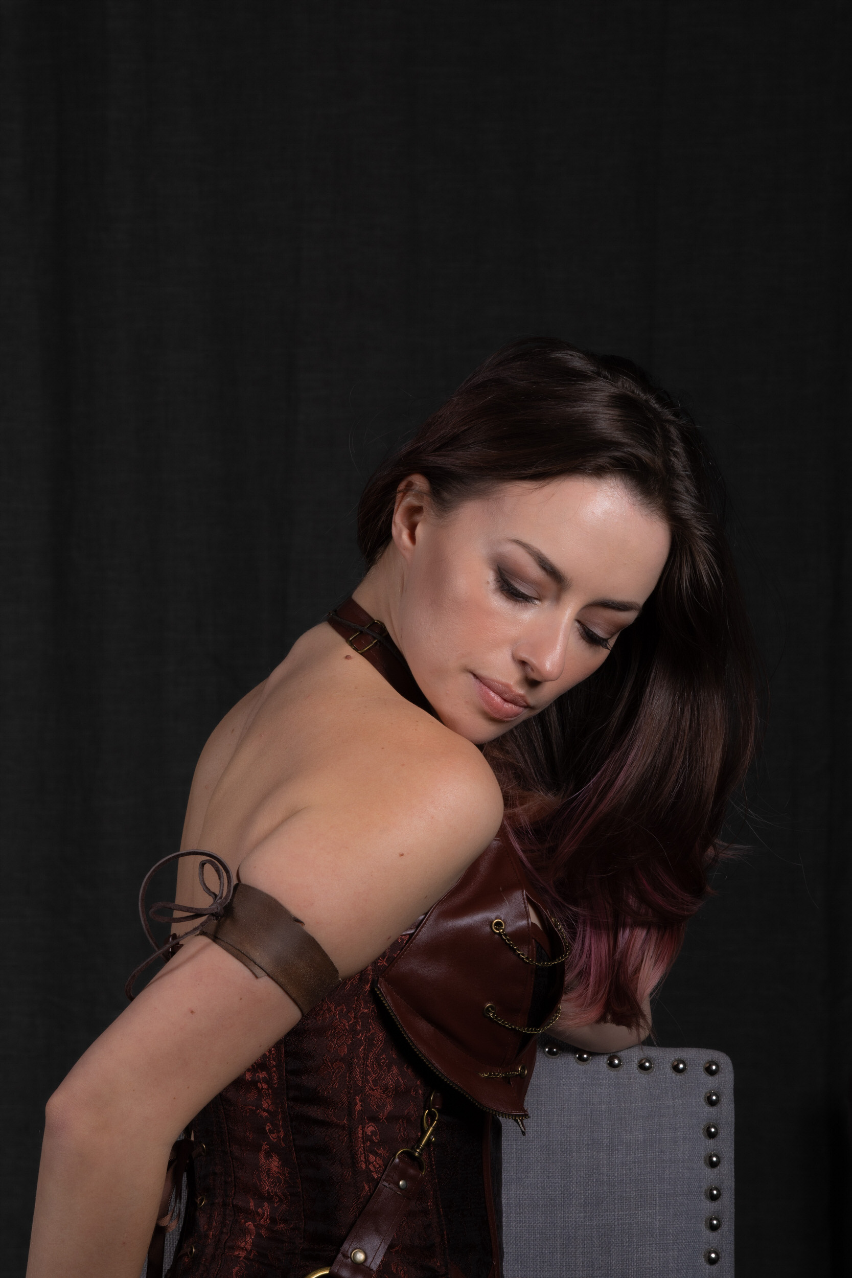 a woman with long, dark hair, styled with a hint of pink, poses against a dark backdrop. she wears an elaborate steampunkinspired corset with leather accents, her left arm adorned with a matching leather bracer. the focus is on her delicate features and the intricate details of her attire, creating a captivating blend of beauty and edginess.