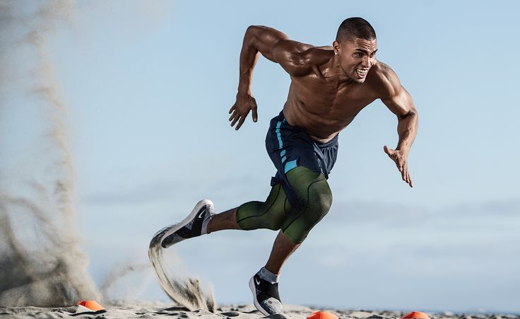 a muscular man wearing athletic shorts and compression tights is sprinting across a sandy beach. he is focused and determined, with his arms outstretched and his legs pumping hard. he is training hard to improve his speed and endurance.