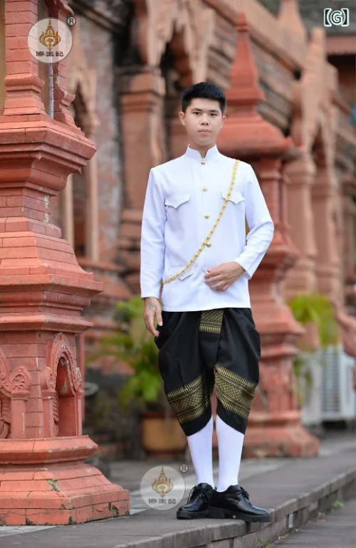 a young man dressed in traditional thai clothing, wearing a white shirt with gold buttons and a black and gold patterned skirt, stands in front of a red brick building.  he is smiling and wearing black leather shoes and white socks.