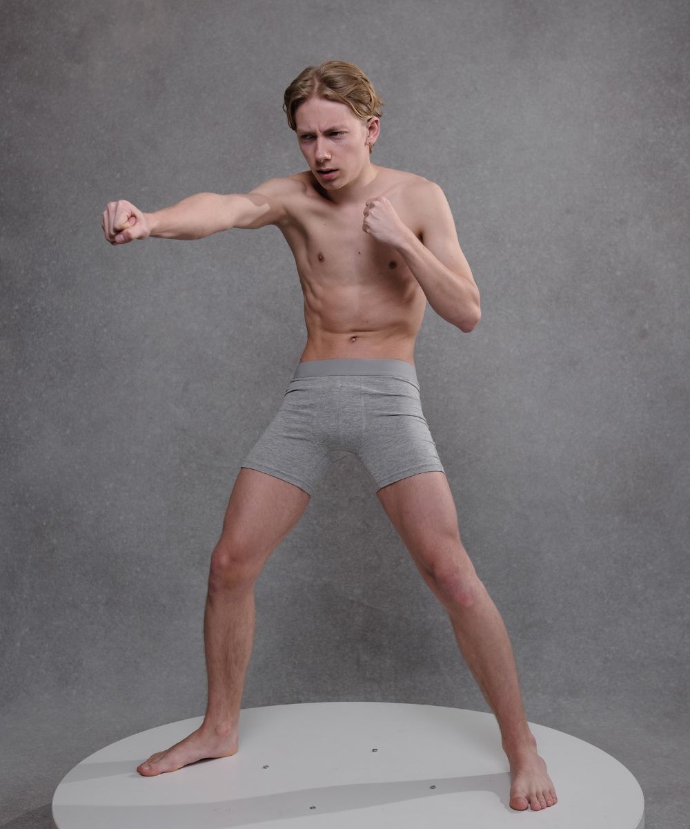 a photo of a young, slender man with a lean physique. he is shirtless and wearing grey boxer shorts. the man is in a fighting stance, with his fists clenched, and is positioned on a white circular platform against a muted grey background. the overall impression is one of fitness and athleticism.