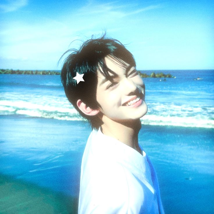 a young asian man with short dark hair is smiling broadly and looking off to the side. he has a white star sticker on his hair and is wearing a white tshirt. the background is a bright blue ocean with waves and white sand beach. the image is taken outdoors in the daytime.