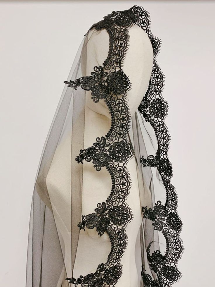 closeup view of a black wedding veil displayed on a mannequin. the veil is made of sheer black tulle with a substantial border of intricate black lace. the lace features floral and scrolllike designs. the overall aesthetic is elegant and gothic, suitable for a dark or alternative wedding theme.  the image focuses on the detail and texture of the lace and tulle.