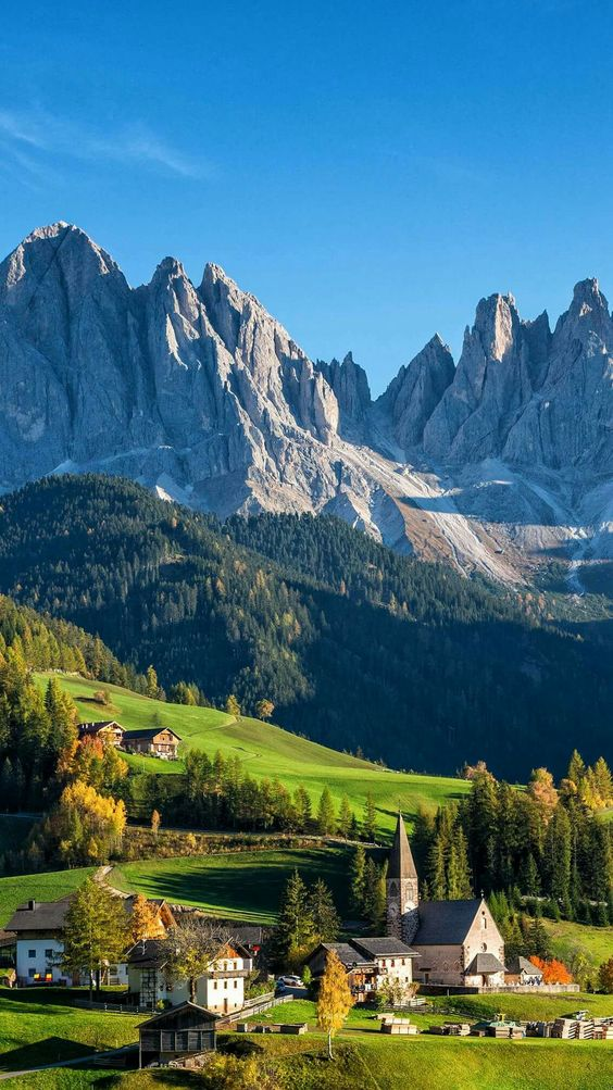 a picturesque village nestled amidst rolling green hills and forests, with the majestic dolomite mountains towering in the background. the village features traditional houses and a prominent church steeple, all bathed in warm sunlight.