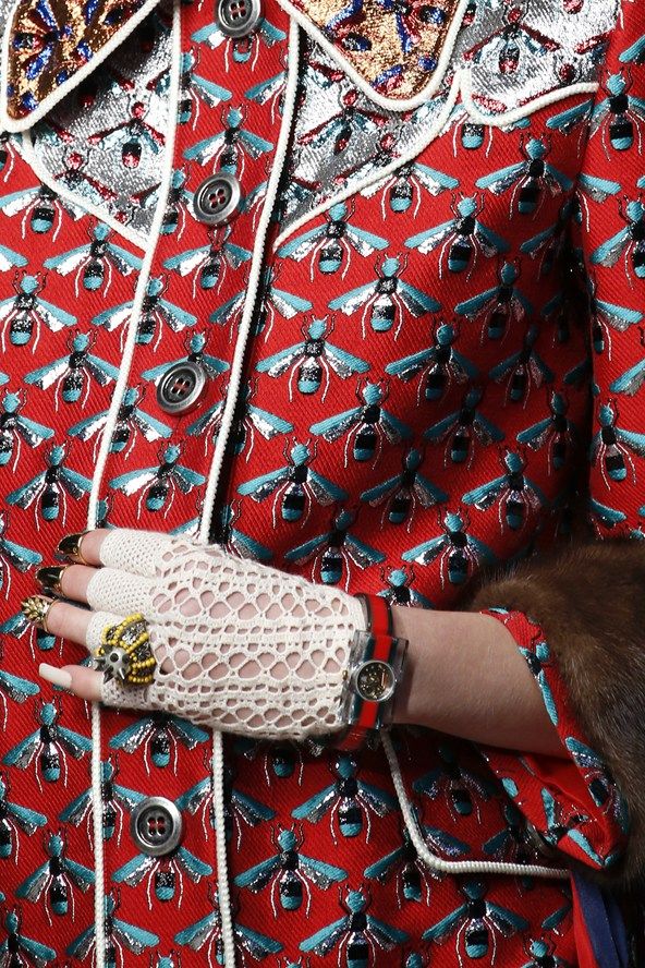 a closeup shot of a woman's hand wearing a white lace glove and a gucci watch.  the hand is resting on a red jacket with an intricate embroidered pattern of bees in gold and silver threads. the jacket features a westernstyle collar and detailing. the overall image showcases the details of the luxurious gucci clothing and accessories.