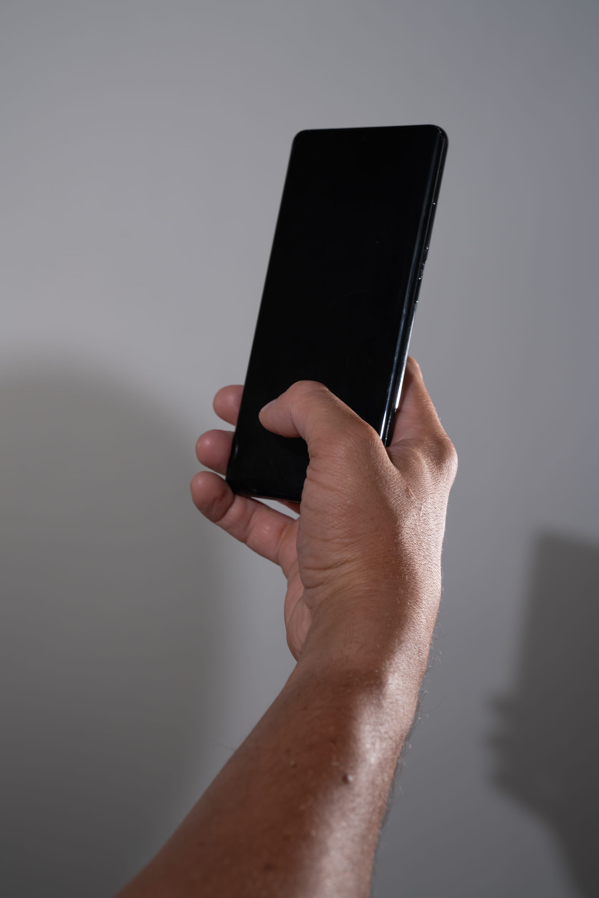 a closeup shot of a person's hand holding a black smartphone against a white wall. the screen of the phone is blank. the hand and arm are illuminated by natural light, casting a shadow on the wall.