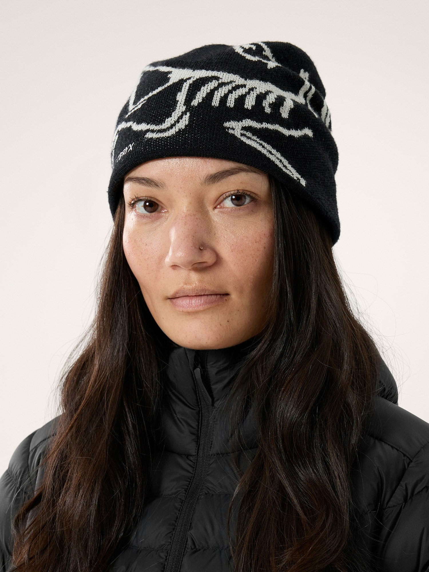 closeup shot of a woman with long brown hair wearing a black knitted beanie with a white skull graphic. she is also wearing a black puffer jacket. the background is a plain light gray. the image is welllit and shows the details of the beanie and the woman's face clearly.