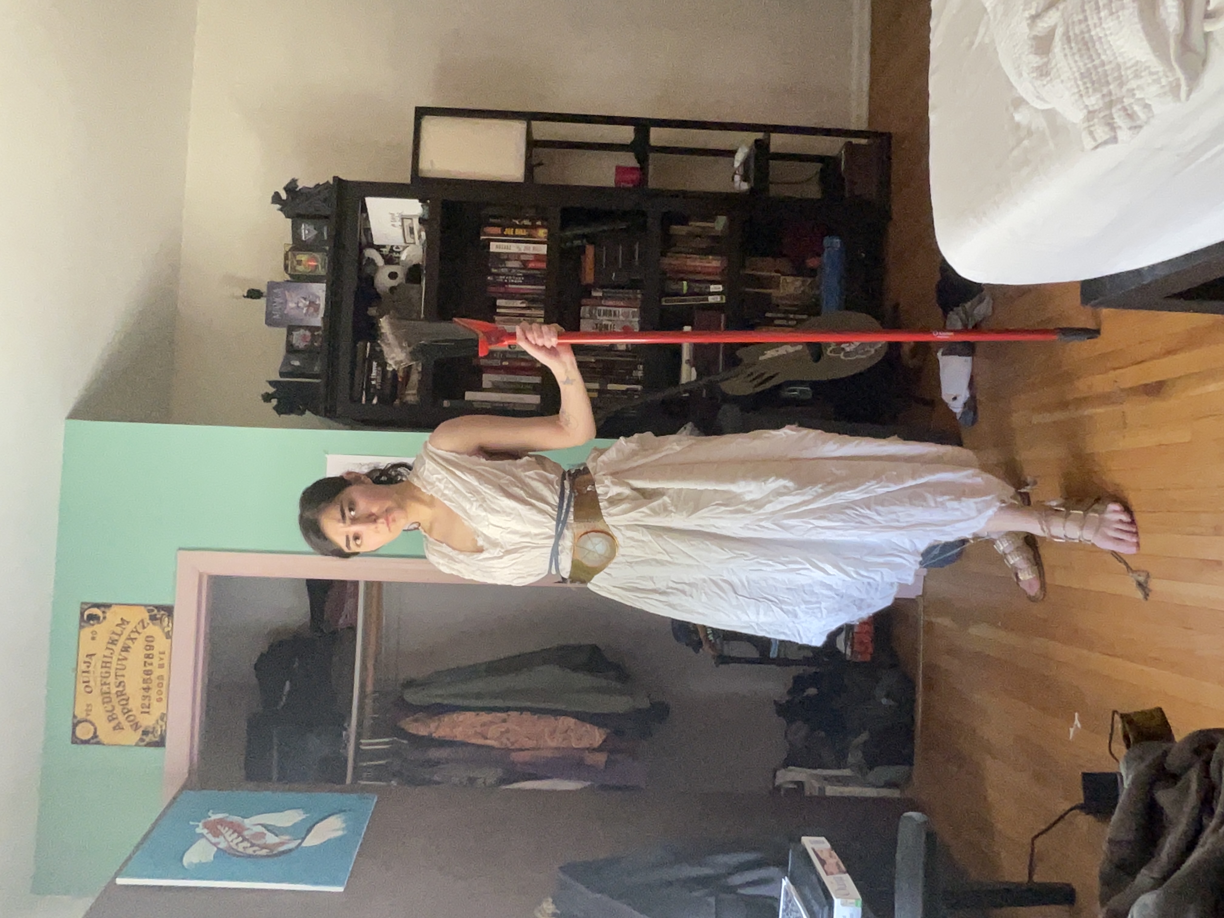 a woman dressed in a white dress with a belt and sandals stands in a bedroom holding a red staff. there is a closet behind her with an ouija board above it, a bookshelf to her right, and a bed to her right.