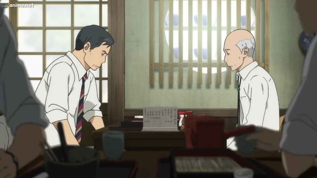 two men are sitting opposite each other at a low table in a japanese restaurant. the younger man has short dark hair and wears a white shirt and striped tie. the older man is bald with gray hair on the sides and wears a white shirt with a light blue tie. a menu sits on the table between them. the room has tatami mat flooring and a shoji screen window that lets in soft natural light, creating a calm and peaceful atmosphere.