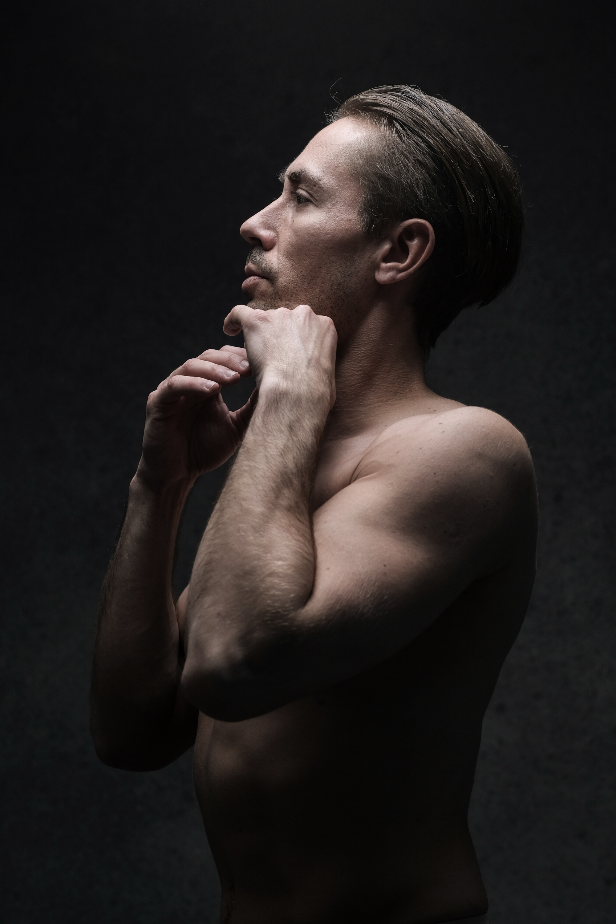 a shirtless man stands against a dark gray background. he is in profile, gazing upward with a contemplative expression, his hands gently touching his face. the dramatic lighting illuminates his muscular physique and creates a moody and introspective atmosphere.