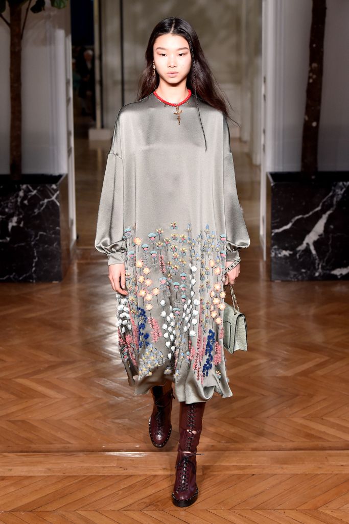 full shot of a young asian female model showcasing a longsleeved, gray embroidered dress with floral accents. the dress falls to her midcalf and features delicate embroidery. she's wearing dark burgundy high laceup boots. a small, lightcolored handbag is slung over her shoulder. her long black hair is straight, and she sports a cross necklace.  the background is a classic runway setting with marble and polished wooden flooring.  the overall impression is one of elegant, understated autumnal fashion.