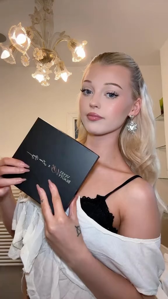 a woman with blonde hair is holding a black makeup palette in front of her. she is wearing a black lace top under a white blouse. she has heartshaped earrings and a tattoo on her forearm. there is a chandelier in the background.