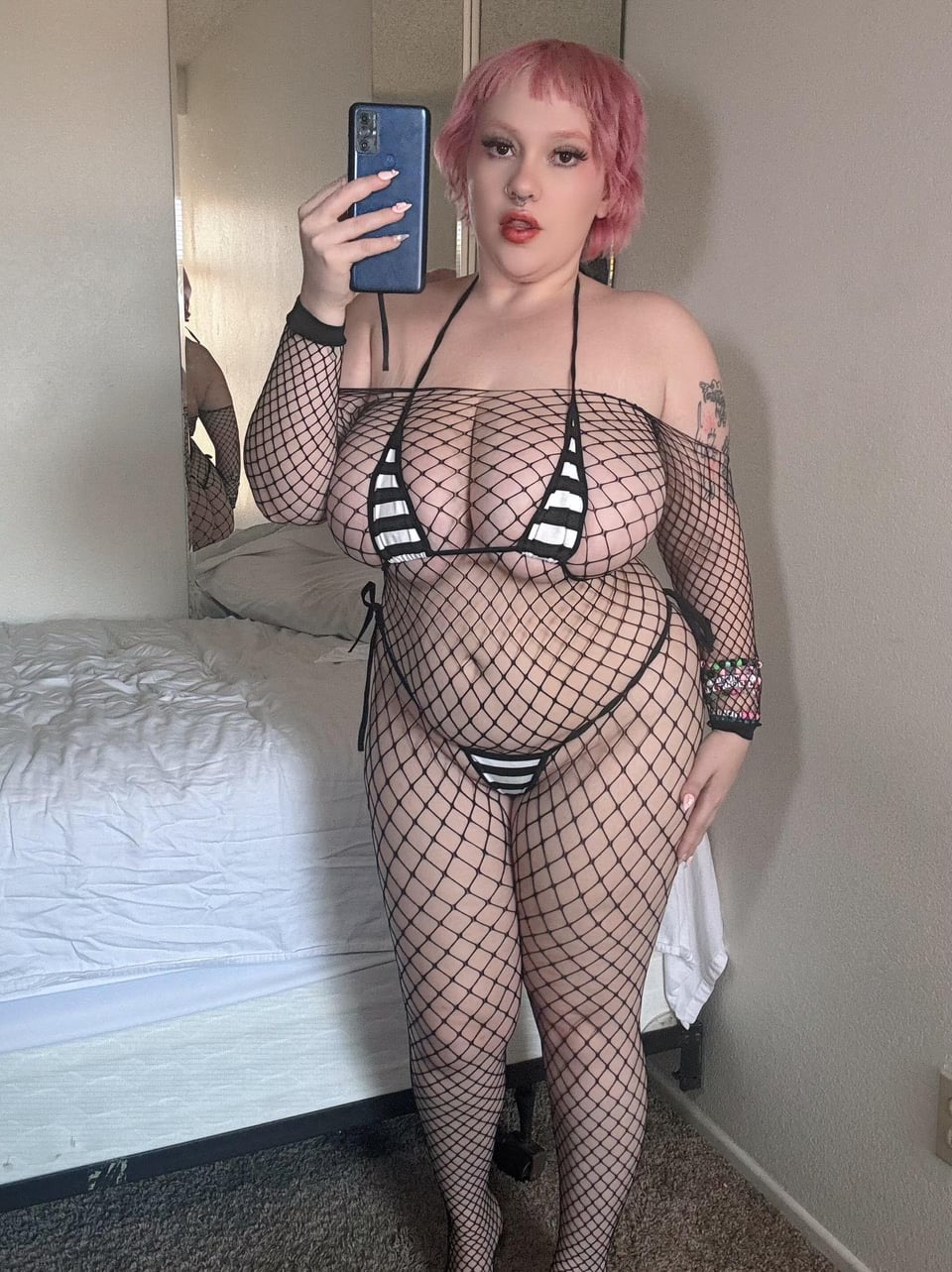 a woman with pink hair is taking a selfie in a bedroom. she is wearing black fishnet lingerie with a black and white striped bikini underneath. the room has a bed with white sheets and a mirror. the woman has tattoos and a body positive attitude. she is holding a phone in her left hand.
