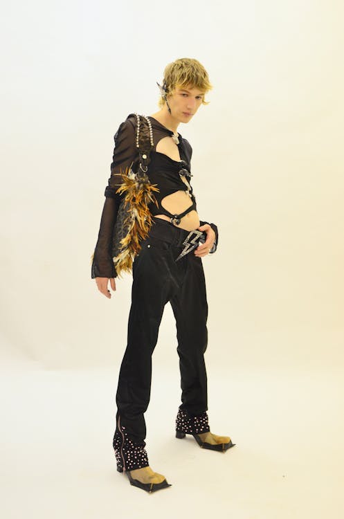 a man stands in a fashion shoot. he wears a black, longsleeved sheer top with a black cutout harness top. he is wearing black pants with a silver lightning bolt belt buckle. he is wearing studded boots with tan pointed tips. he has a bag with fur accents.