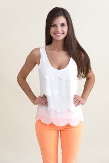 a young woman with long brown hair is smiling and looking at the camera. she is wearing a white sleeveless top with a scalloped hem and orange coral colored pants. the top has a white to pink ombre effect. the background is a light beige wall.