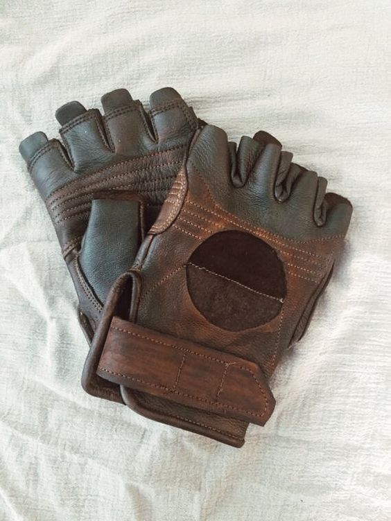 a pair of brown leather gloves with a wrist strap, designed for weightlifting or other gym activities.  they are made of brown leather and have a stitched design. they are lying on a white fabric background.