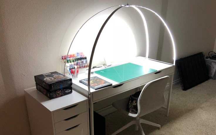 an art desk with a curved led light stands next to a drawer unit in a room. there is a chair in front of the desk. the desk has a green cutting mat on top and is also holding containers of paints, markers, game boxes, and a dvd case.