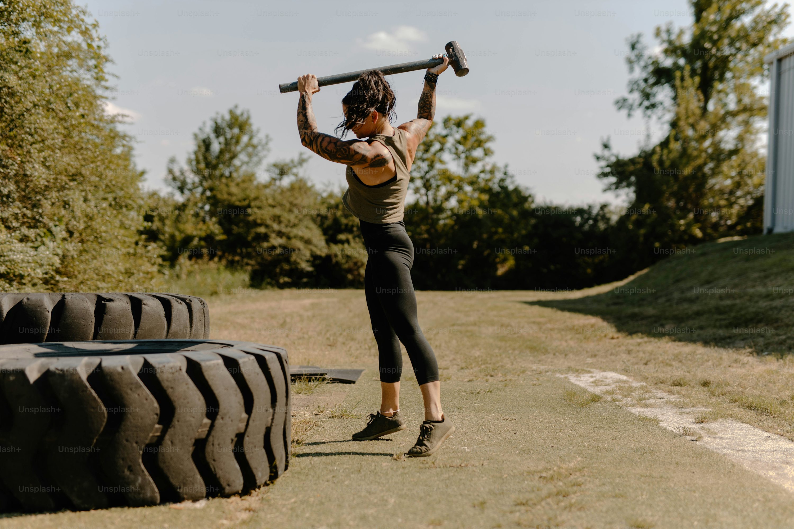 the image shows a strong, tattooed woman performing a tire flip exercise outdoors.  she is using a sledgehammer to hit a large tire that rests on the ground. she's wearing athletic clothing—leggings and a green top— and has her hair tied back. the setting appears to be a grassy area with trees in the background, suggesting an outdoor gym or training space. the overall feel of the image is one of strength, determination, and athleticism.