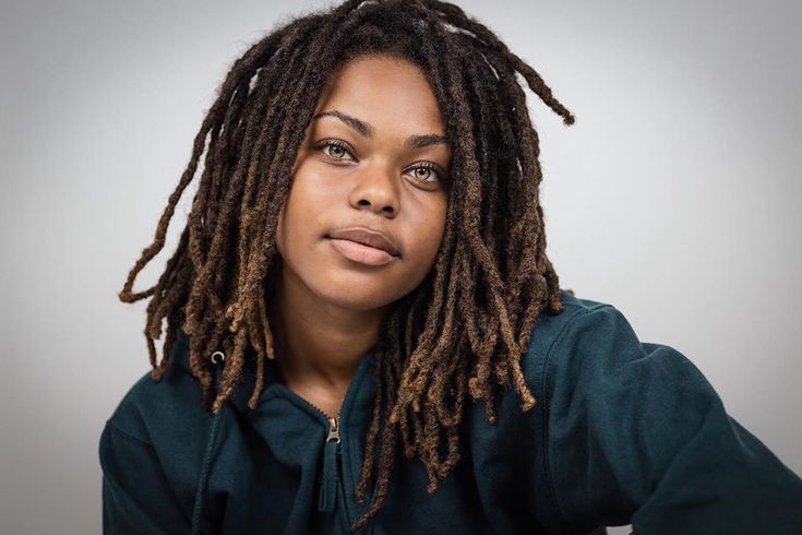 a portrait of a woman with dreadlocks, wearing a dark blue hoodie. she has hazel eyes and a neutral expression. the photo appears to be taken in a studio with natural light.