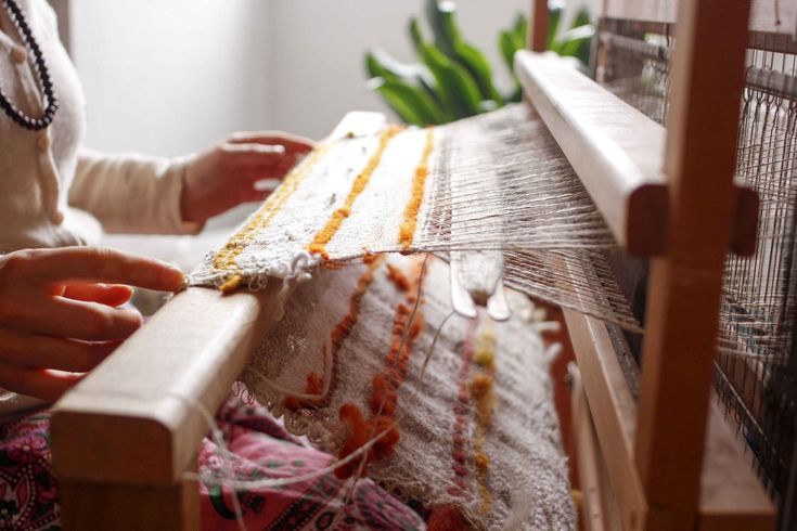 a closeup shot captures a person weaving on a wooden loom. their hands are visible as they manipulate the yarn and fabric. the textile being created features a variety of colors and textures. the loom itself is prominently displayed, showcasing its structure and the intricate process of weaving.