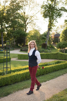 a man with long blonde hair is walking along a garden path. he's wearing a stylish outfit: a dark vest over a white shirt and red pants. he has a single red rose in his hand. the setting is a beautiful park with lush green hedges and trees, bathed in the warm sunlight of a summer day. the overall mood is romantic and elegant.