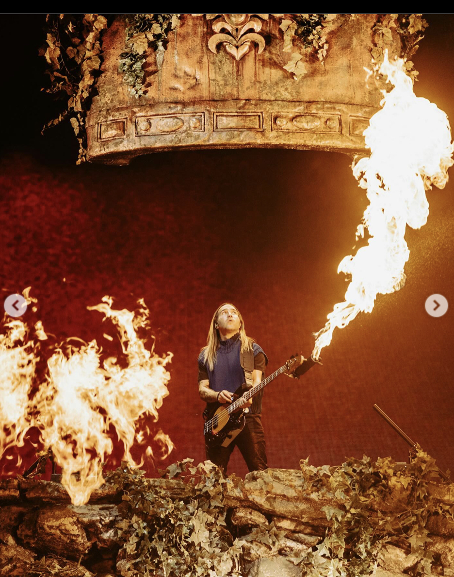 a man in a blue shirt and jeans plays a bass guitar on stage while engulfed in flames. he is looking up at the flames as if they are a spectacle he's enjoying. the image is taken at a concert where the stage is lit up by the flames and there is smoke coming from the flames. the man is surrounded by logs and green leaves which suggests the stage is designed to be a forest setting.