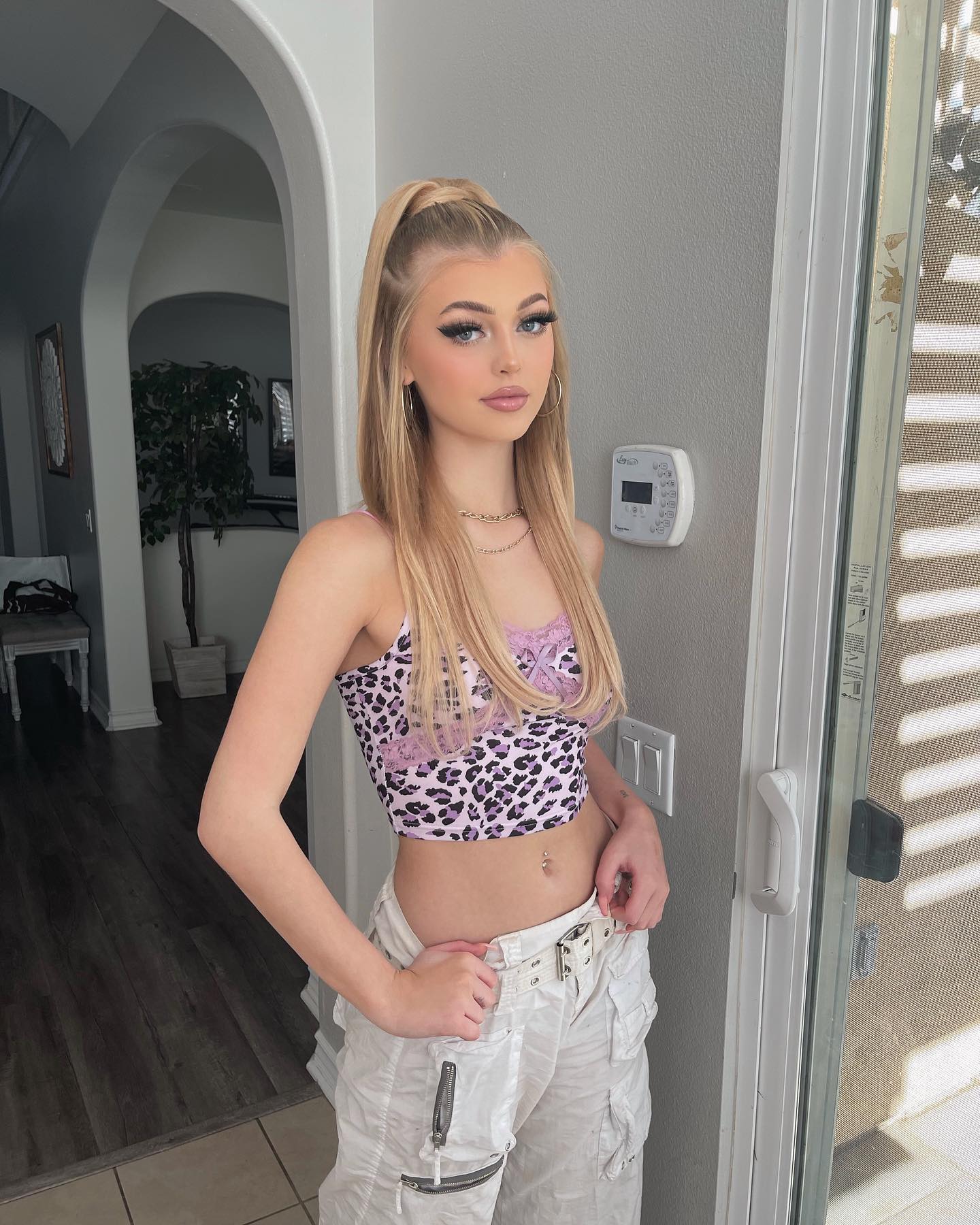 a young woman with blonde hair in a high ponytail stands indoors near a doorway. she's wearing a leopard print crop top with a purple lace trim and loosefitting white cargo pants. she is also wearing gold hoop earrings and several gold necklaces. her hands are on her waist. the background includes a hallway with a visible living area and a closed glass door. a thermostat and light switch are mounted on the gray wall behind her.