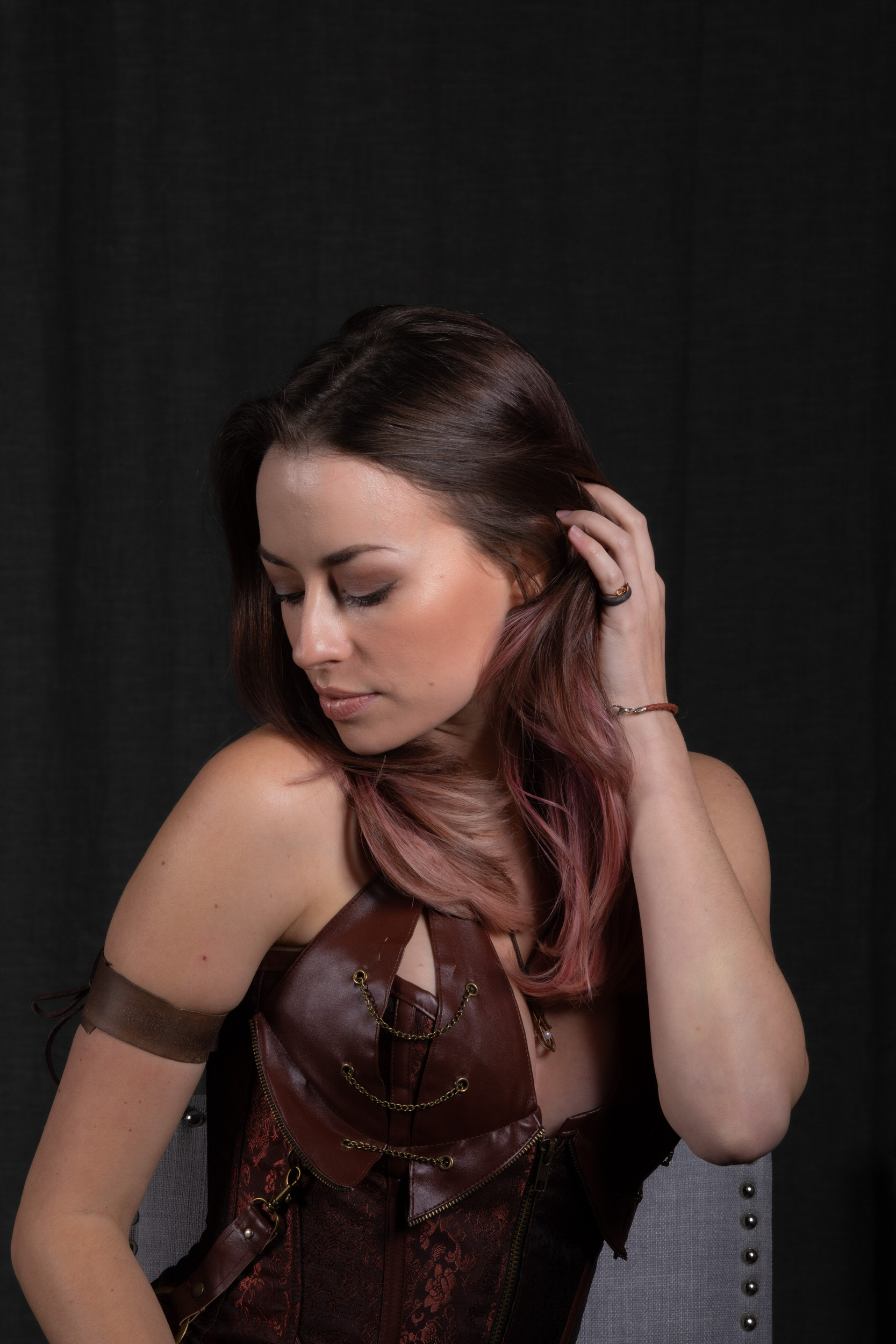 a young woman with pinkhighlighted hair poses against a black backdrop. she wears a brown steampunkinspired corset adorned with buckles and chains, and a matching arm cuff. her gaze is directed downwards, and she gently holds a strand of hair with her hand. the image has a soft, studiolit aesthetic, emphasizing the details of her outfit and features.